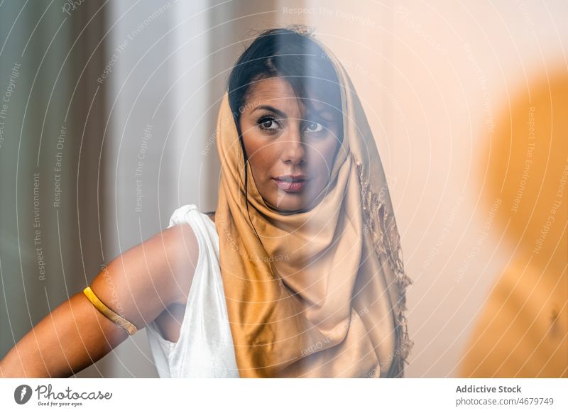 Through glass of Muslim woman near window appearance traditional authentic culture hijab feminine style room ethnic muslim headwear headdress home light design