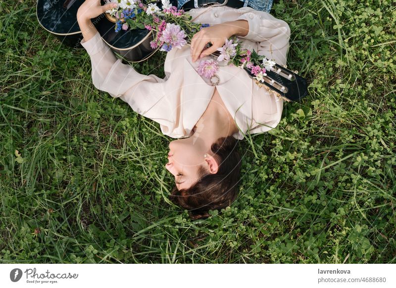 Young woman lying on the grass holding guitar covered with flowers outdoor musician person playing instrument strumming lifestyle song sound entertainment