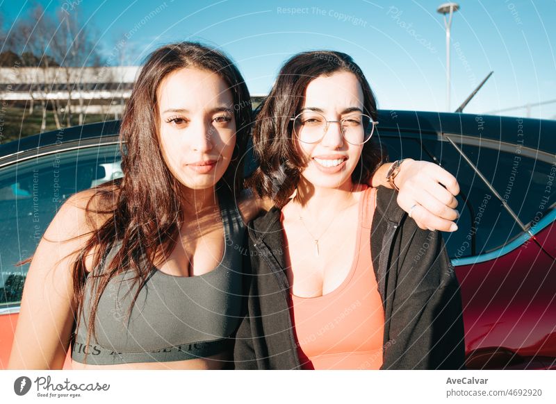 Two young sporty woman friends serious to camera before joining the gym, resting against car. Training losing weight with friends together smiling to camera happy. Friendship between sisters concept