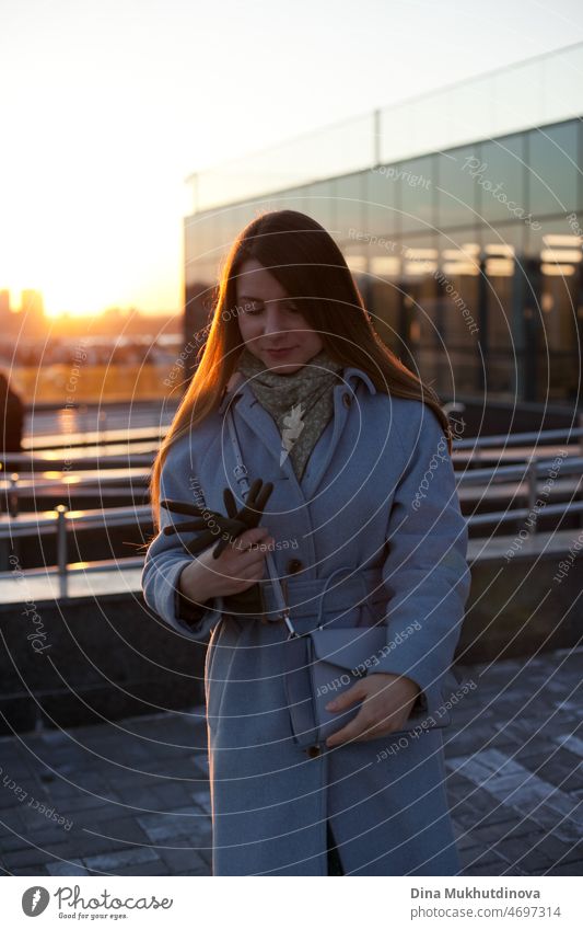 Woman in stylish blue coat leaving office at sunset. Beautiful attractive young woman walking on the street at sunset or sunrise early in the morning near modern architecture buildings in the city. Casual candid lifestyle.