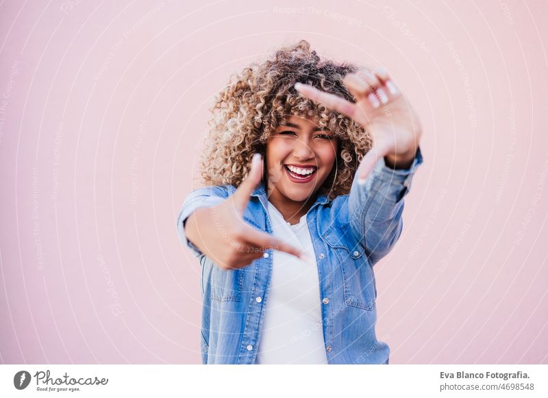 portrait of beautiful hispanic woman afro hair outdoors in spring doing frame with hands. pink wall happy smiling city casual clothing lifestyle curly hair