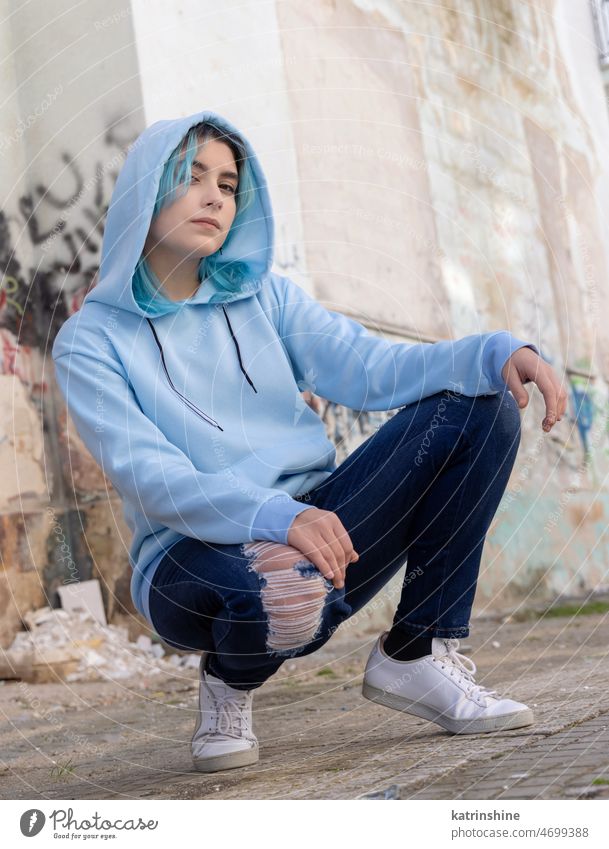 Blue haired Teenage girl in light blue oversize hoodie sitting on her haunches near graffiti wall Teenager mockup jeans blue haired teen girl outdoors urban