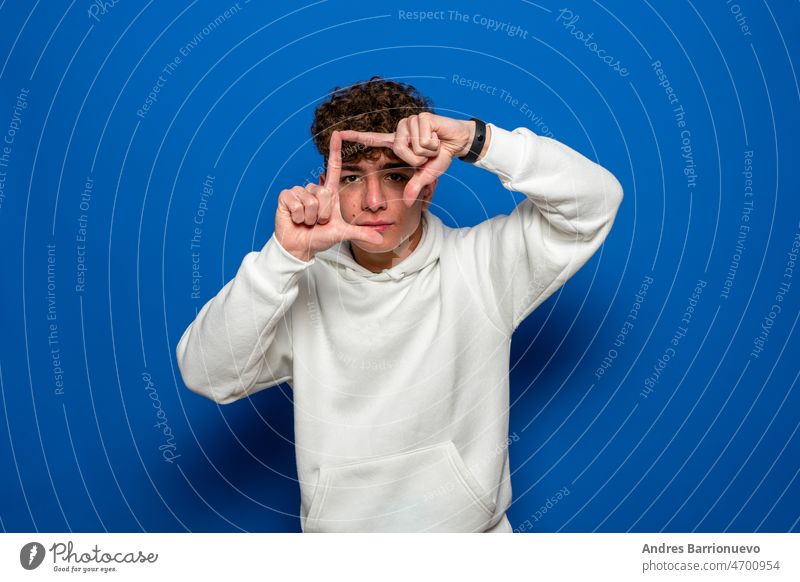 Young handsome brunette man with curly hair wearing white sweatshirt over blue background smiling making frame with hands and fingers with happy face. Concept of creativity and photography.