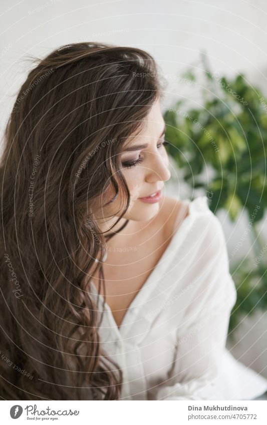 Beautiful young attractive brunette woman with long brown hair  in white shirt, candid lifestyle photo near window with natural morning light with romantic mood. Woman in apartment with green tropical plants as background, minimal interior home.