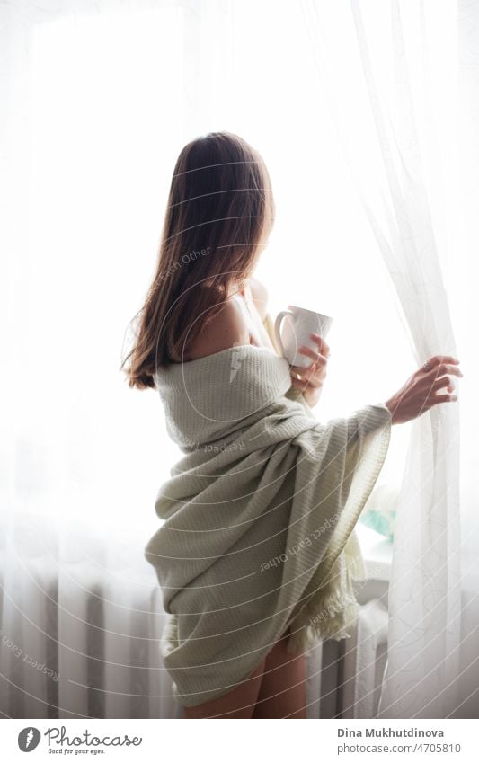 Young beautiful brunette woman from behind in cozy blanket  in the morning at her home. Apartment living millennial lifestyle drinking coffee. comfort