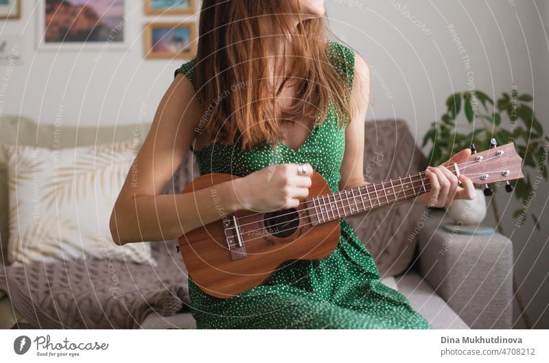 Young brunette woman in green dress playing ukulele at home. Apartment living. Musician talented person playing small guitar as hobby or profession. beautiful