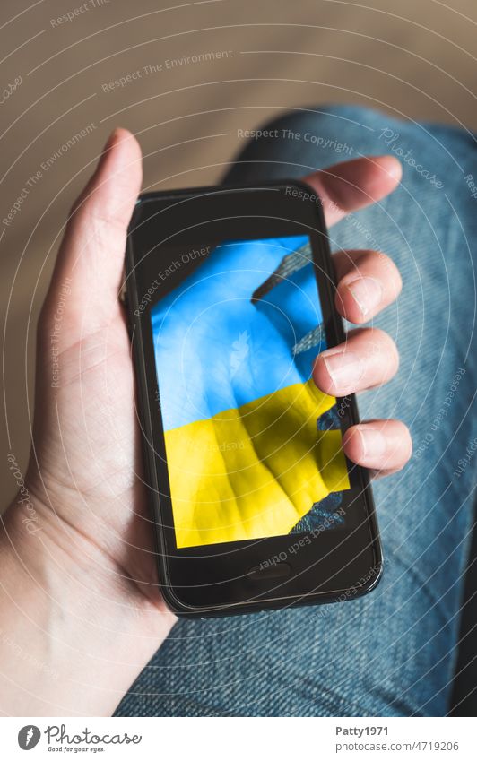 Close-up of a hand holding a smartphone, on the display of which the hand holding it, painted in Ukrainian national colors, can be seen translucent Hand