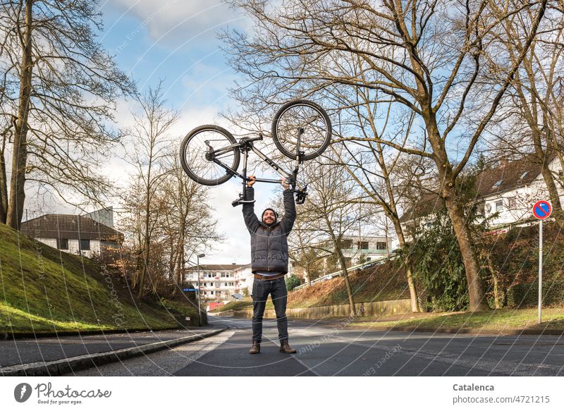 Young man lifts his bike cyclists urban Gray daylight Day trees Residential area Action Cycling Sports Town Winter Bicycle Street person Sky Clouds Force Green