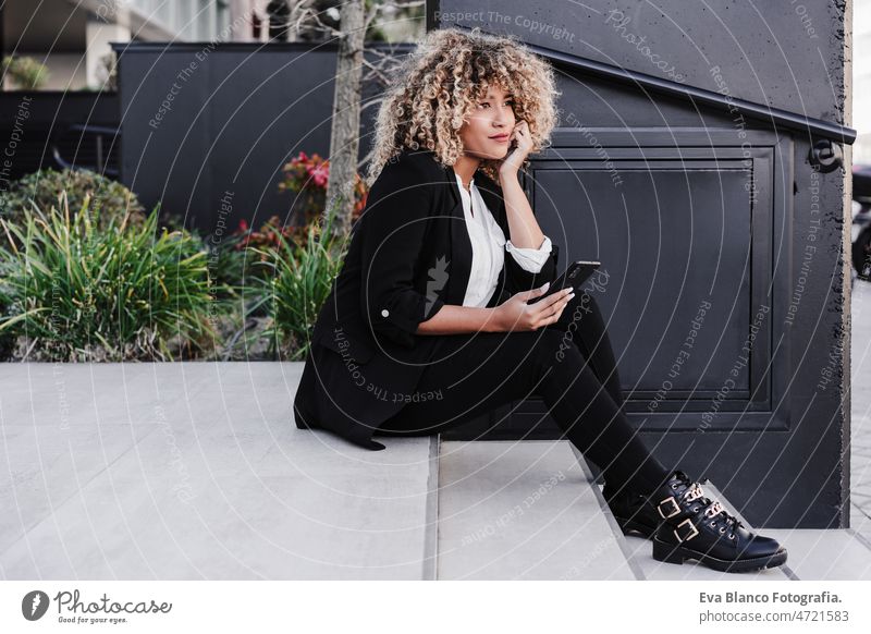 beautiful smiling business woman using mobile sitting on stairs in city. Buildings background afro hispanic mobile phone skyscraper building young curly hair