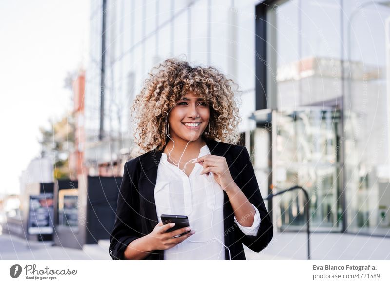 beautiful smiling business woman using mobile phone and headphones in city. Buildings background afro hispanic skyscraper building young curly hair music