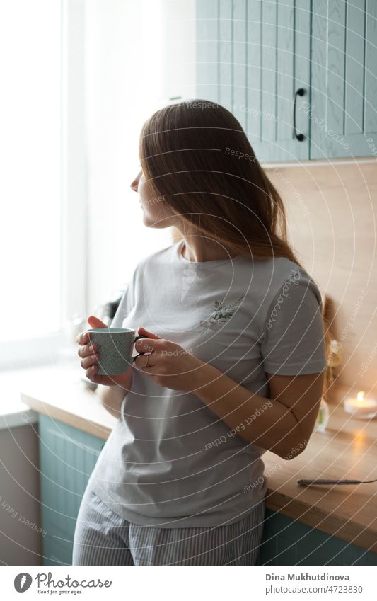 young woman at home drinking coffee in the morning from ceramic cup, smiling to new day standing in the kitchen and looking to the window good morming candid