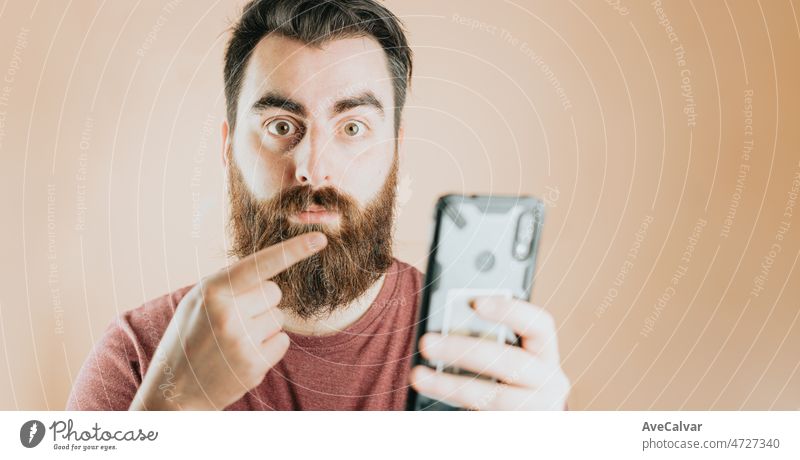 Young bearded handsome hipster man surprised pointing his smart phone, amazing news, unexpected reaction. Soft tone color background, expression of normal people. Technology and connections