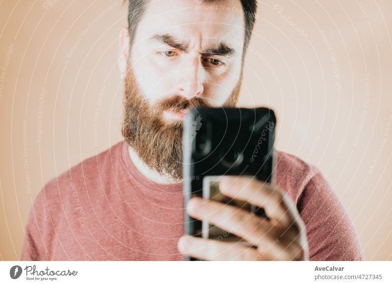 Young bearded handsome hipster man serious worried after reading his smart phone, amazing news, unexpected reaction. Soft tone color background, expression of normal people. Technology and connections