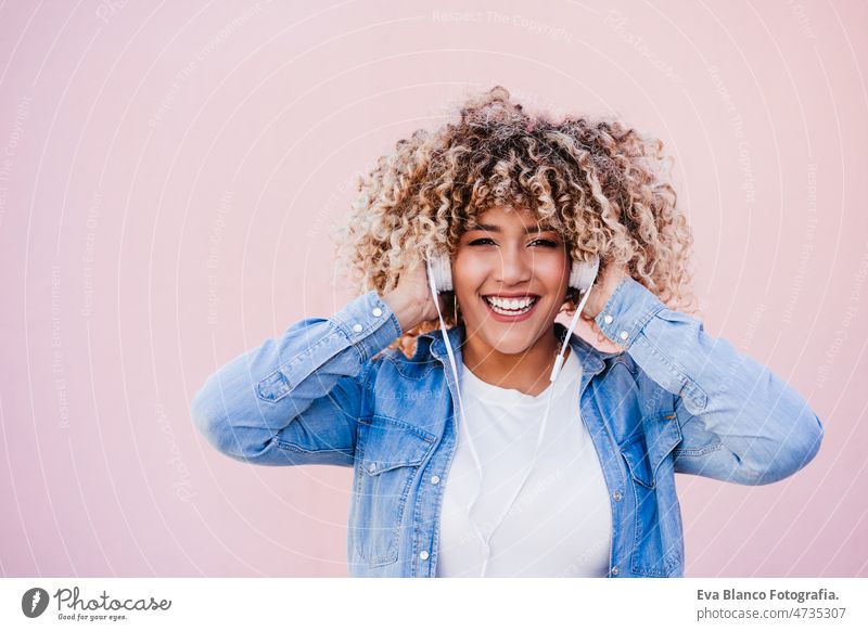 portrait of smiling hispanic woman with afro hair in city using mobile phone and headset. lifestyle listening body positivity curvy selfie technology internet