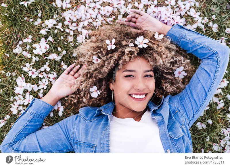 portrait of happy hispanic woman with afro hair lying on grass among pink blossom flowers.Springtime spring close up almond tree colorful curly hair lifestyle