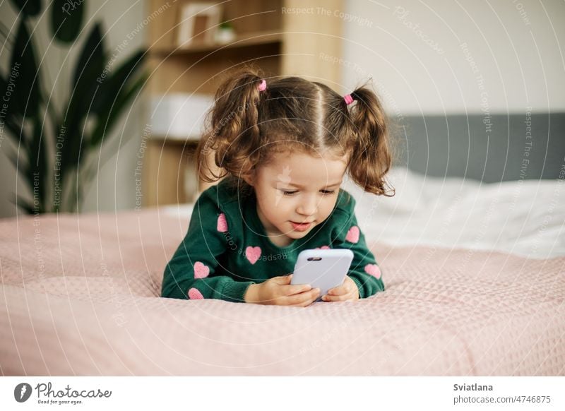 A little girl is happily looking at a smartphone, playing a game or watching cartoons child development cute smile mobile kid bed caucasian touch happy