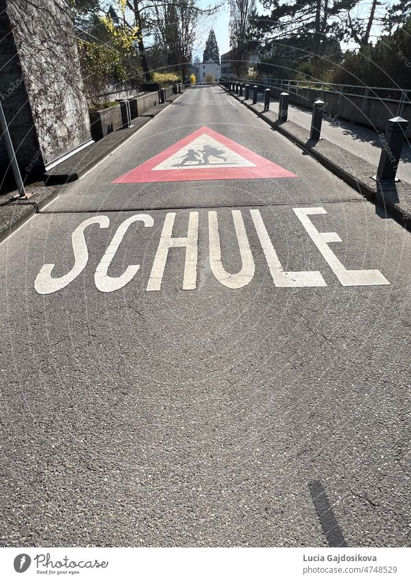 Warning sign of the way to school in German language  in Switzerland. Road sign is red triangle with running children in white field and text Schule, which means School.