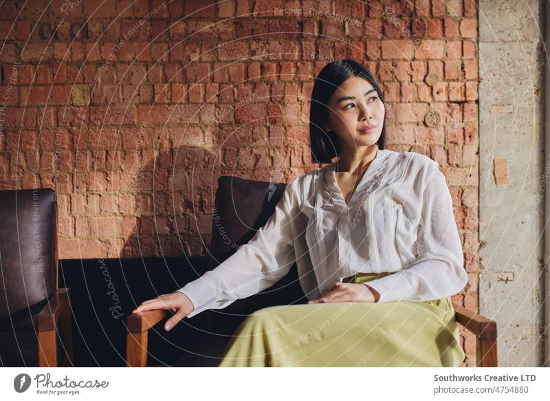 Portrait of mid adult Chinese businesswoman sitting on chair in front of exposed brick wall and looking away portrait chinese retro asian one people indoors day