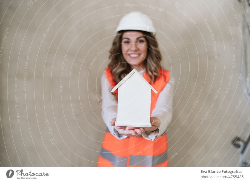 professional confident architect woman in construction site holding house model. Home renovation blueprints workspace protective helmet protective jacket