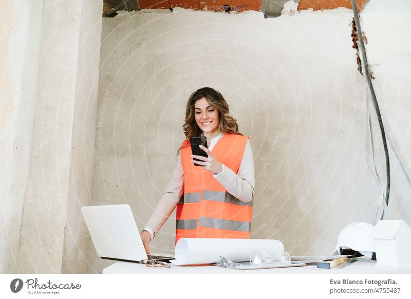 professional confident architect woman in construction site using mobile phone holding blueprints workspace protective helmet protective jacket real estate