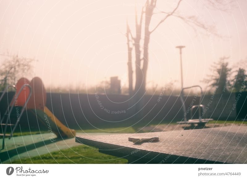 A table tennis bat is lying on a table tennis table, in the background a playground can be seen. Playground Table tennis Table tennis table Table tennis bat