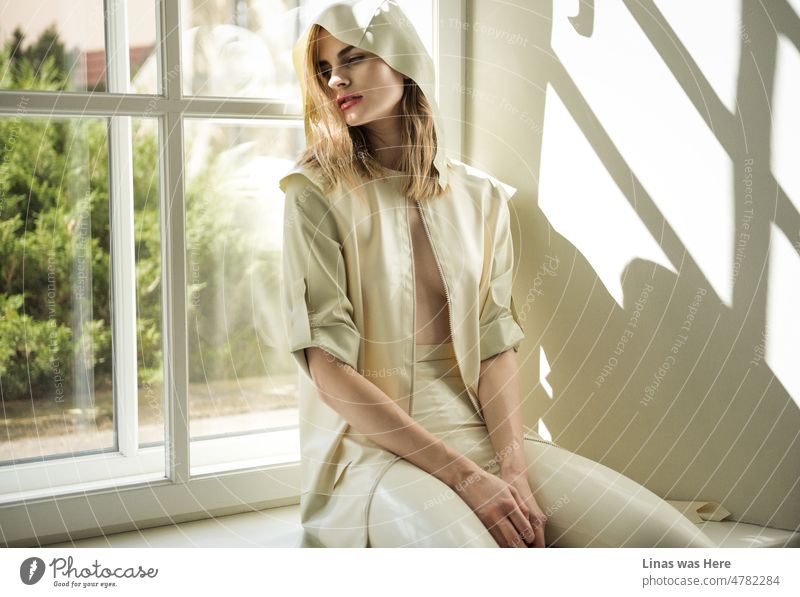 A gorgeous fashion model dressed in a white latex raincoat is sitting next to a sunny window. It’s springtime though the girl feels moody. A perfectly beautiful woman and the rebirth of nature in this one.