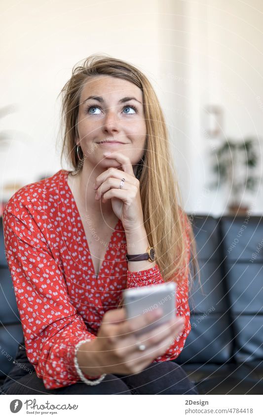 Portrait of single beautiful Caucasian woman hold cellphone in hand and sitting on sofa while looking away thoughtfully thinking smartphone couch person mobile