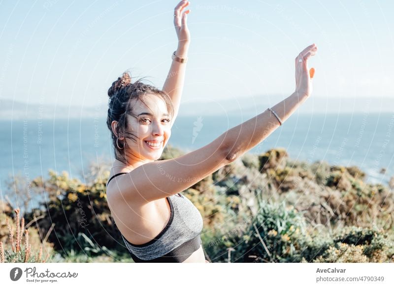 Young woman celebrates finishing a outdoor exercise session on the coastline. Dancing and raising arms to the air. Happy young people training during a sunny summer day preparing for the beach body