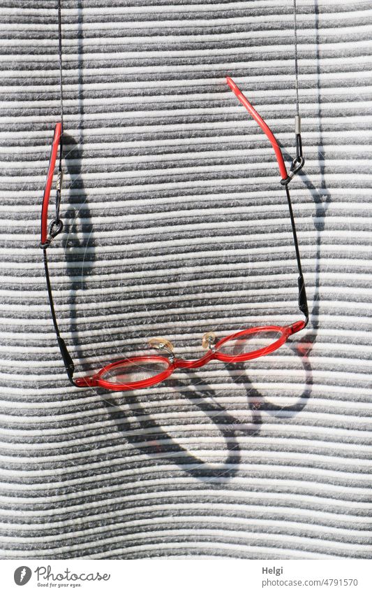 Order of glasses on ribbon - red glasses with big shadow hanging on glasses ribbon in front of gray and white striped sweater | UT Frühlingslandluft Eyeglasses