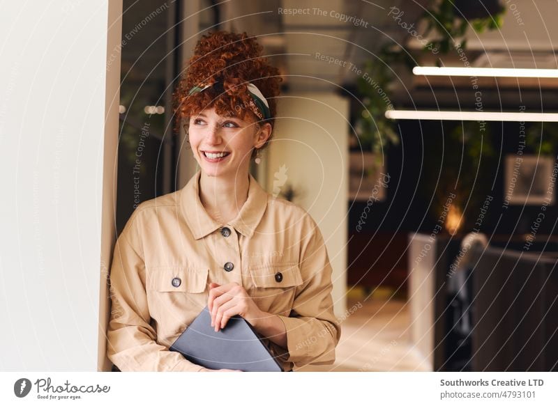 Portrait of confident young white woman with red curly hair smiling and looking away holding laptop in coworking space portrait smile happy office indoors day