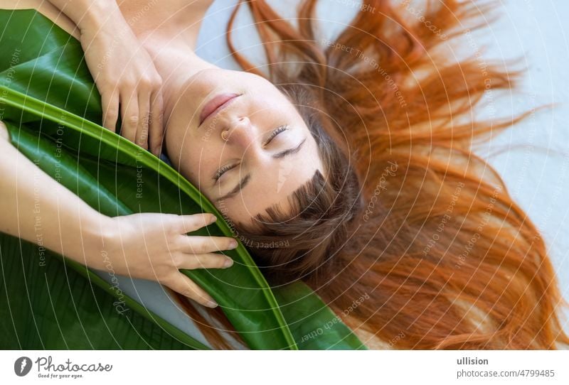 seductive sensual portrait a young sexy woman with red hair loves nature and nestles on green decorative banana leaf, lying on the gray studio floor copy space