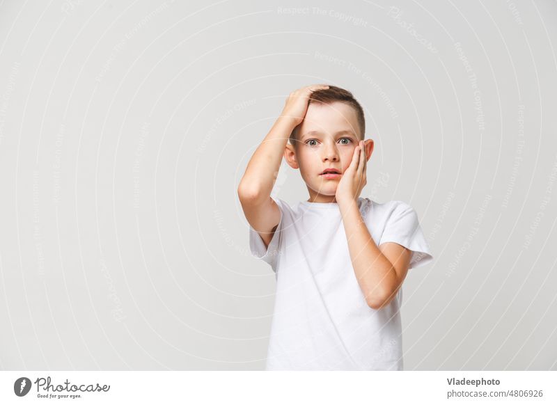 Terrified boy in white t-shirt with both hands on head on gray