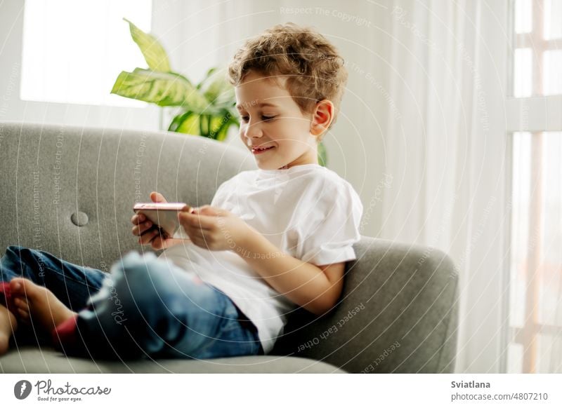 A cheerful boy with a phone in his hands is sitting in a chair and playing a game or watching cartoons. smartphone little home window cute internet technology