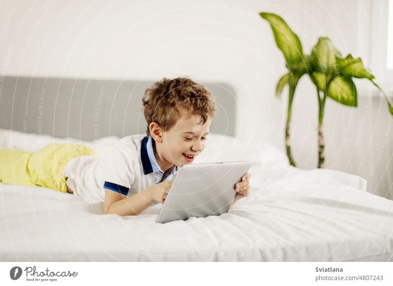Curly-haired boy is lying on the bed in the bedroom and playing computer games on a tablet lies technology happy child indoors kid one internet fun smiling