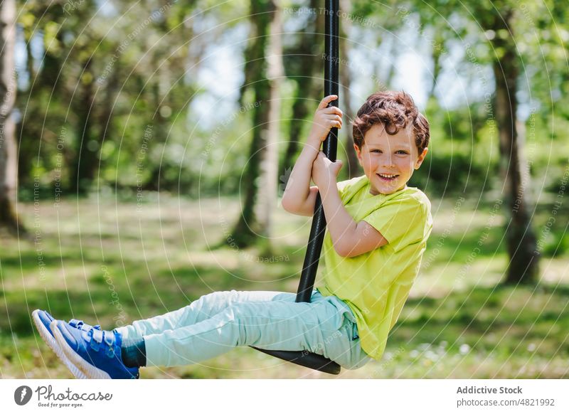 Cheerful little kid on swing rope on playground smiling at camera child smile childhood cheerful activity boy park positive portrait hang excited joy summer