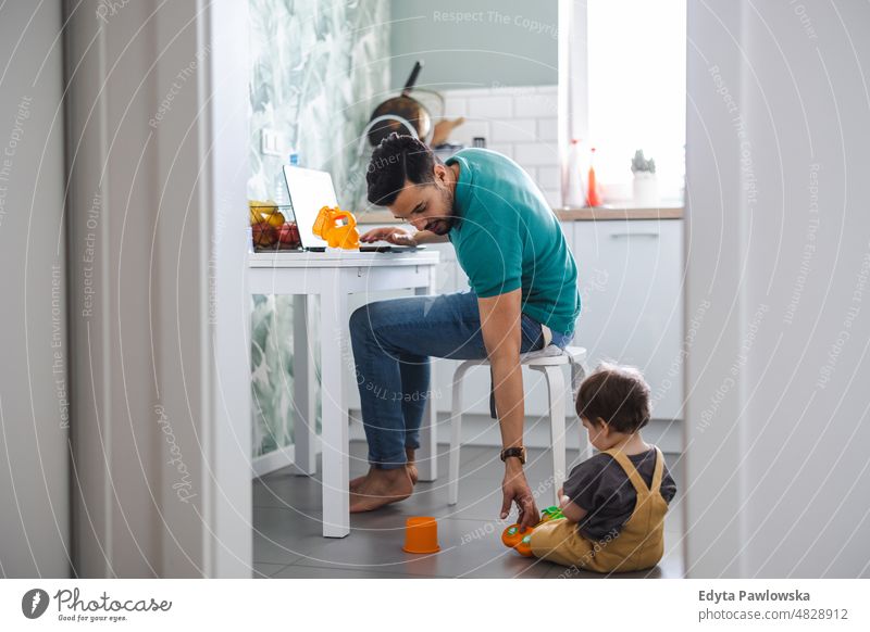 Father working on laptop while child playing on kitchen floor adult affection baby bonding boy candid care childhood domestic enjoying family fun happiness