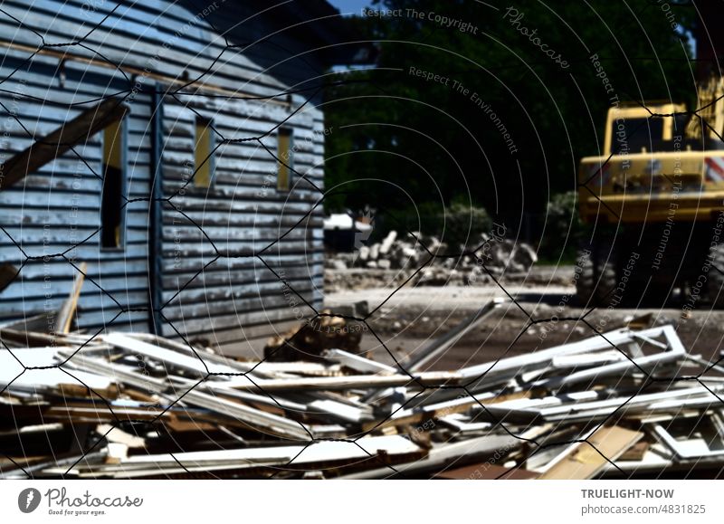 Behind a light wire fence a demolition lot: there are wall remains, old beams and boards lying around in front of a dirty blue facade and an old yellow excavator is also still there