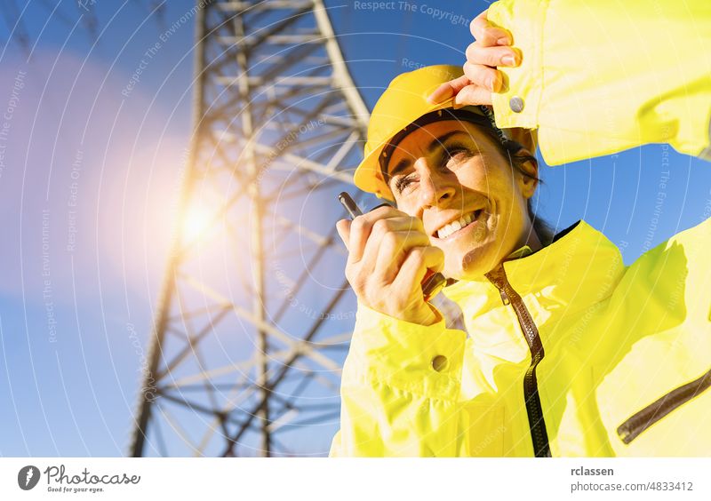 Happy female Electrical engineer talking in a walkie talkie to control a high voltage electricity pylon. Electrical power lines and towers with bright sun