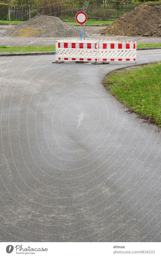 Barriers and a traffic sign - prohibition for vehicles of all kinds - stand on a turning gray asphalt road in front of piles of earth and chippings / construction site / road closure