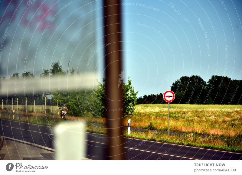 View from bus stop over country road with traffic sign no overtaking and boundary posts to summer field with forest in background and cloudless sky; a cyclist seems to be approaching