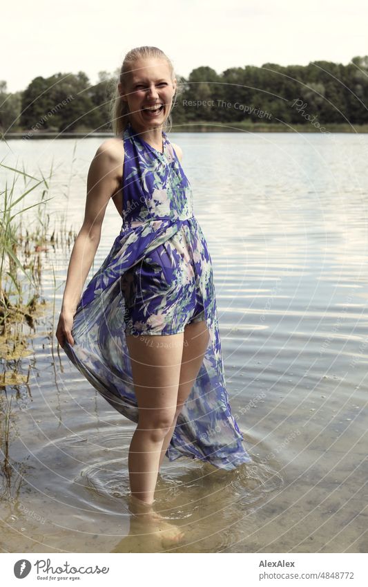 Young red blonde long haired woman stands laughing in dress with feet in lake on shore looking sideways at camera Woman youthful pretty Slim Grass Nature Blonde