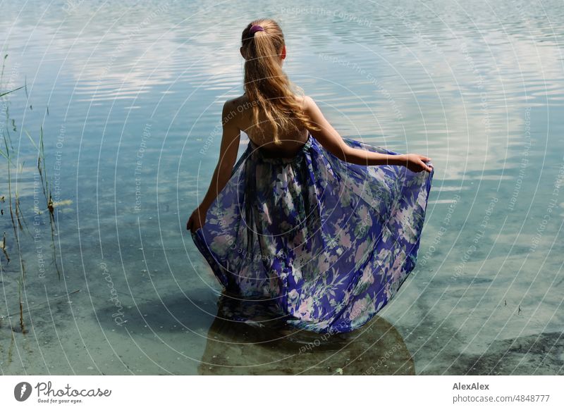 Young red blonde long haired woman standing in dress with feet in lake on shore looking at lake Woman youthful pretty Slim Grass Nature Blonde cheerful