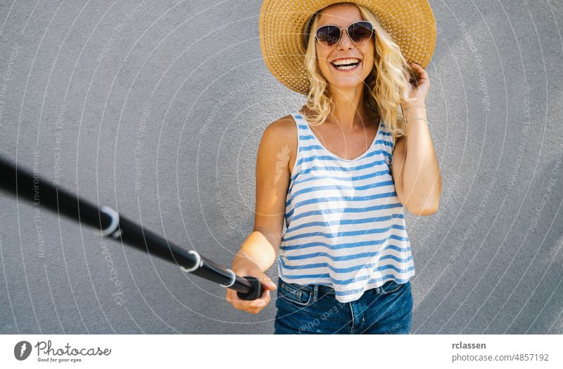 Young girl with straw hat smiling and use selfie stick to take a selfie, on summer in city street. Urban life concept image young hipster trave trip woman smile