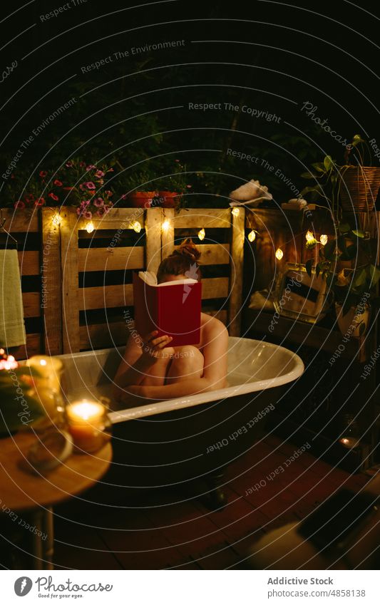 Faceless woman reading book in bath on terrace evening patio hobby pastime naked cover face glow bathtub garland female literature shiny illuminate story novel