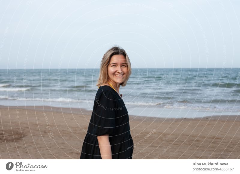 Young woman in casual black dress on the beach, smiling. Horizontal photo of female tourist on the empty beach pretty Beach voyager Woman Ocean Landmark Happy