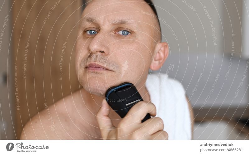 Daily routine male self-care. Handsome young unshaven man with towel on shoulders shaving with modern electric razor trimmer while looking at camera, indoors.