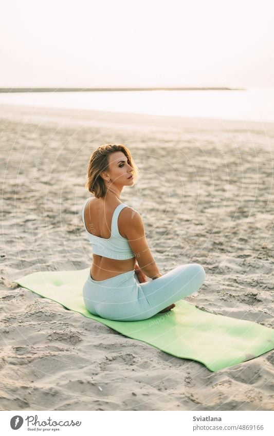 A beautiful girl is sitting on the seashore in the lotus position and resting after yoga or fitness classes. Healthy lifestyle, self-care, activity beach woman