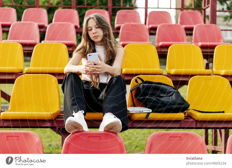 A teenage girl sits on the school bleachers and writes a message on her phone in her free time schoolgirl daytime smartphone stadium texting education sitting