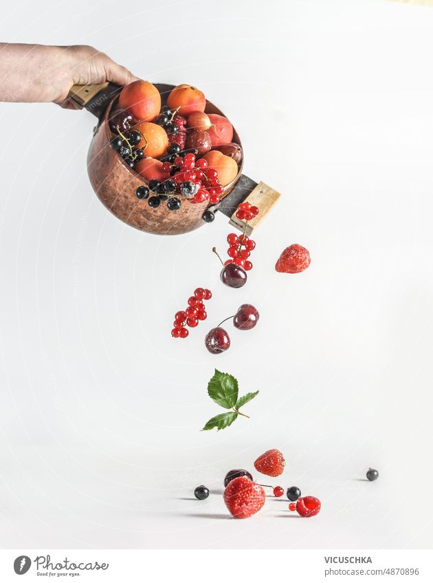 Woman hand holding copper cooking pot with falling summer fruits and berries at white background woman currants strawberries raspberries cherries gooseberries