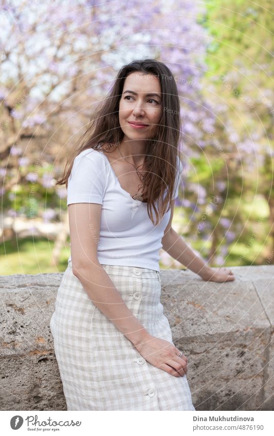 Young female brunette tourist posing near jacaranda tree. Neutral colors in clothes style. Beige color walls. Woman tourist wearing white t-shirt. summer